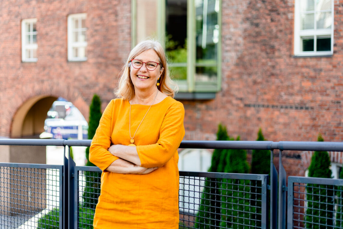 Tarja Muukkonen nojaa rennosti kädet puuskassa ulkona kaiteeseen punatiilisen rakennuksen edessä.