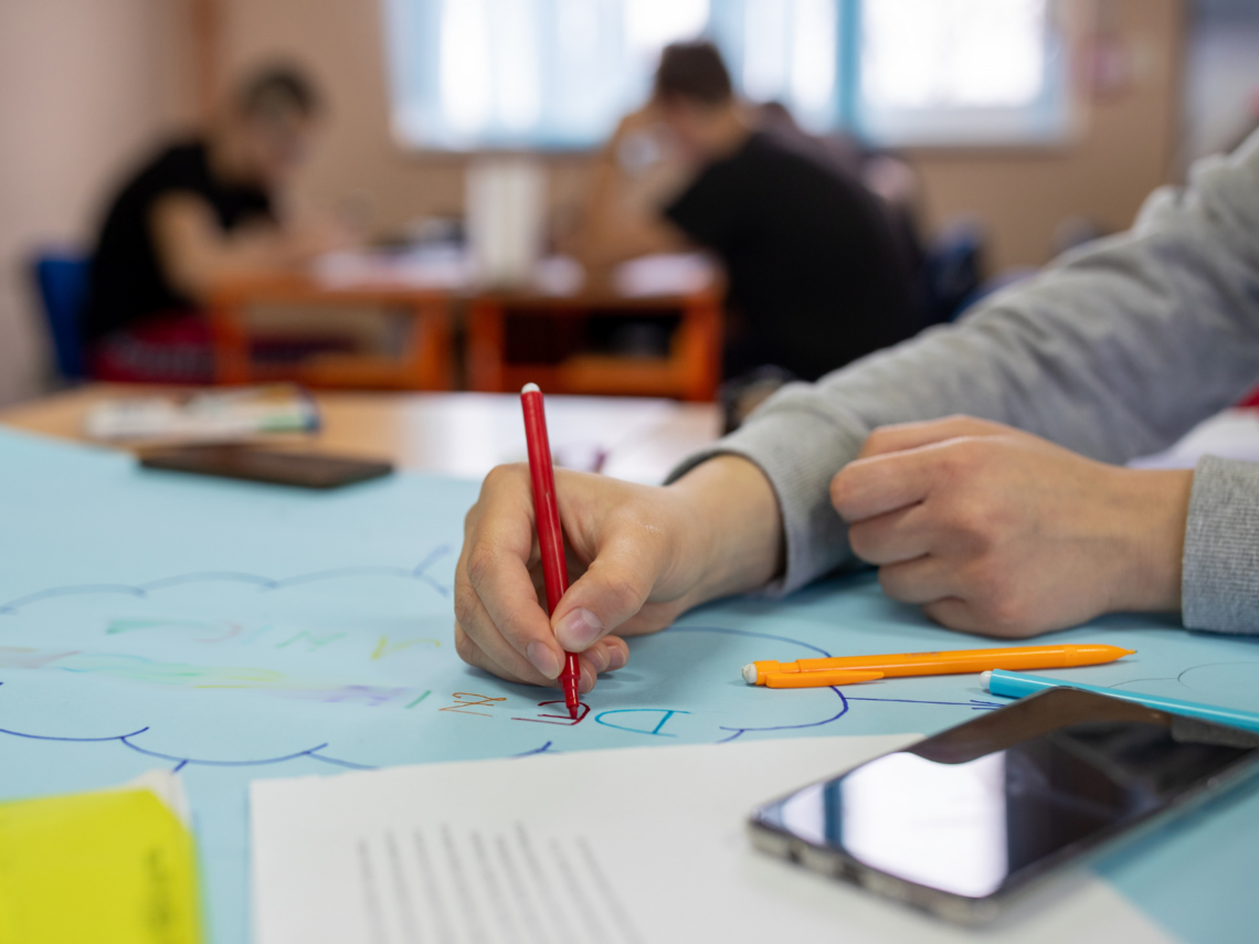 Käsi piirtää värikkäälle paperille tussilla ja kirjoittaa tekstiä. Taustalla näkyy muita työskentelemässä taka-alalla.
