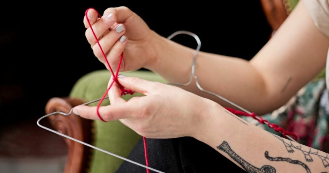 Kädet punovat henkarin ympärille punaista lankaa. Toisessa kädessä on tatuointeja.