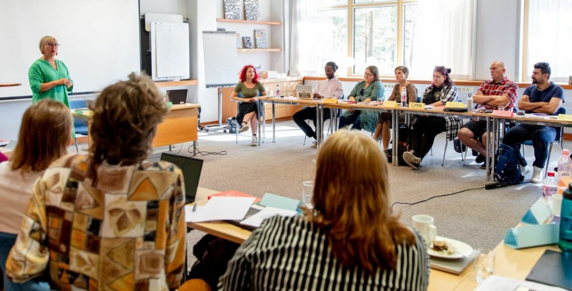 Kymmenen ihmistä istuu valoisassa opetustilassa ympyrämuodostelmassa. Aino-Kaisa Pekonen puhuu ryhmälle.