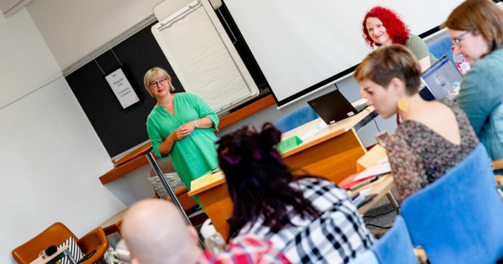 Neljä henkilöä istuu luokkahuoneessa saman pöydän ääressä. Joku puhuu, ja luokan edessä oleva Aino-Kaisa Pekonen ja Tiia Kontkanen kuuntelevat.