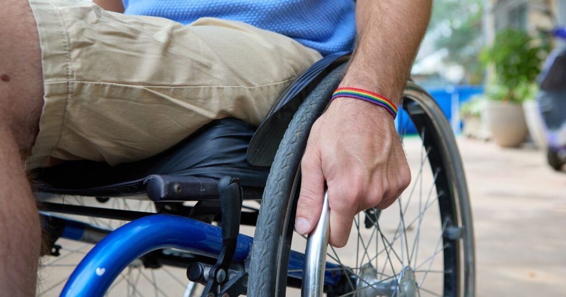 Lähikuva pyörätuolissa istuvasta, jonka käsi pitää kiinni pyörätuolin renkaasta. Ranteessa on sateenkaarinauha.