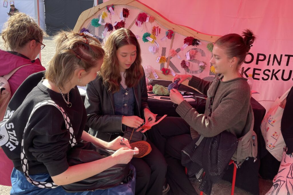 Neljä henkilöä istuu tuoleilla ja tekee tupsuja. Taustalla näkyy kehikko, johon on kiinnitetty tupsuja.