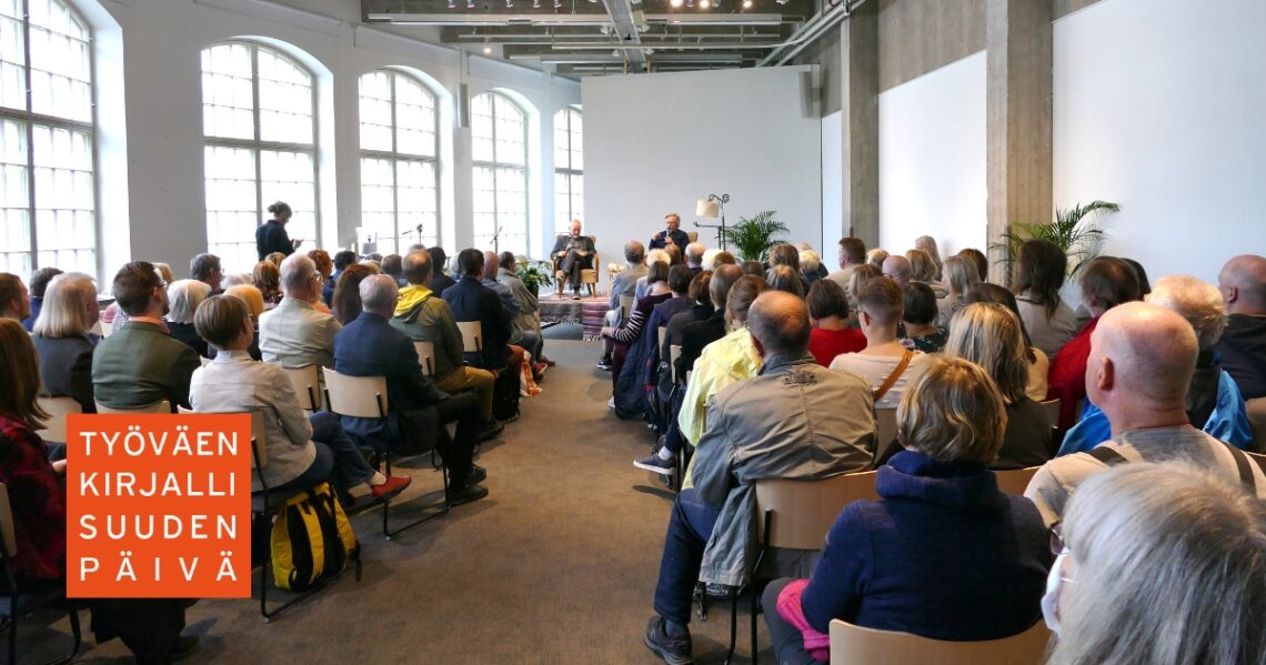 Yleisöä valoisassa salissa kuuntelemassa kahta lavalla keskustelevaa henkilöä. Kuvan päällä lukee Työväenkirjallisuuden päivä.