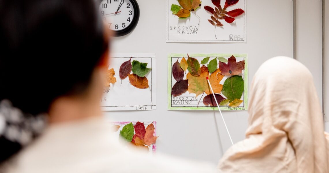 Huiviin pukeutunut osoittaa karttakepillä seinällä olevaa paperia, johon on kiinnitetty syksyn lehtiä. Paperiin on kirjoitettu käsin Syksy on kaunis ja Fatima.