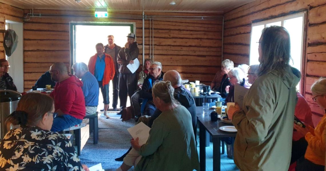 Joukko ihmisiä seisoo ja istuu sisällä mökissä.