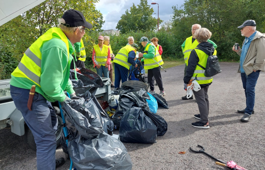 Yksitoista henkilöä peräkärryn luona. Ihmisillä on yllään huomioliivit. Peräkärryn vieressä on täysiä jätesäkkejä ja roskiekeruupihdit.