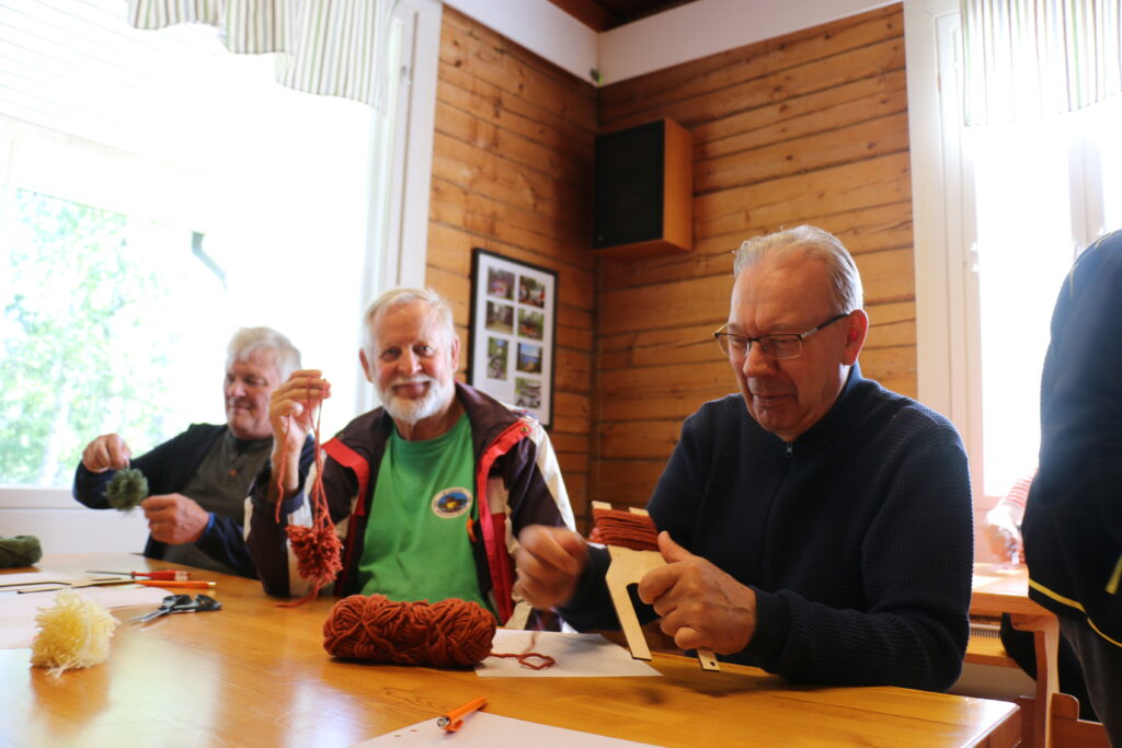 Kolme henkilöä pirtissä pöydän ääressä tekemässä tupsuja langasta.