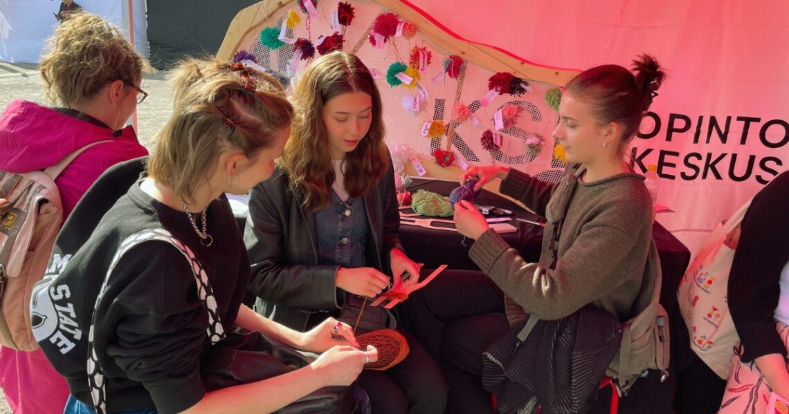 Kolme henkilöä istuu telttakatoksessa ja tekee langasta tupsuja. Taustalla näkyy tupsuja, joita on kiinnitetty kehikkoon.