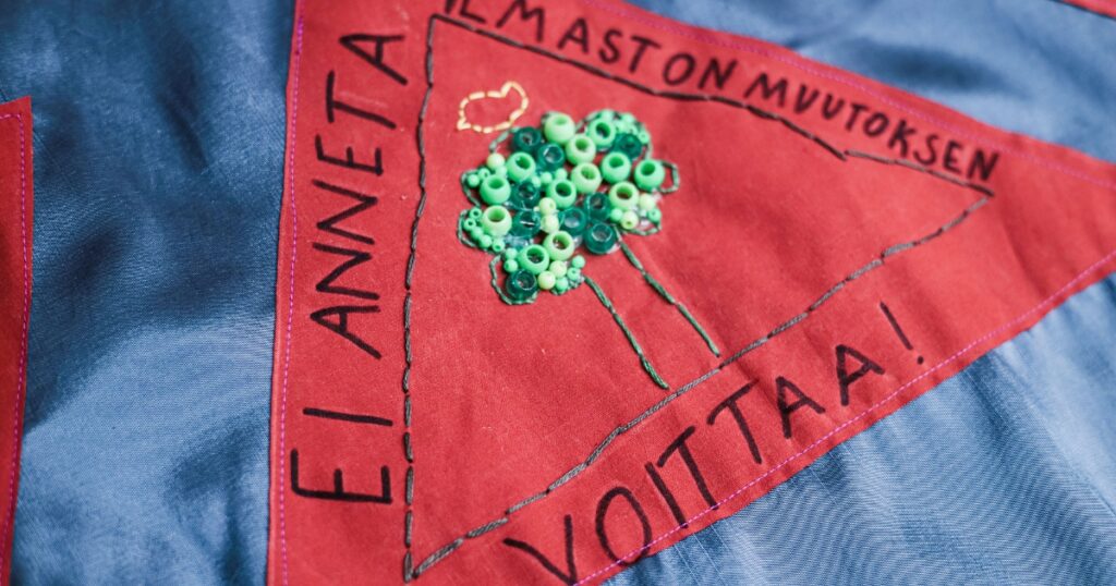 Kolmion muotoinen kangas. Siihen on kirjailtu koristeellinen puu ja kirjoitettu teksti: Ei anneta ilmastonmuutoksen voittaa.