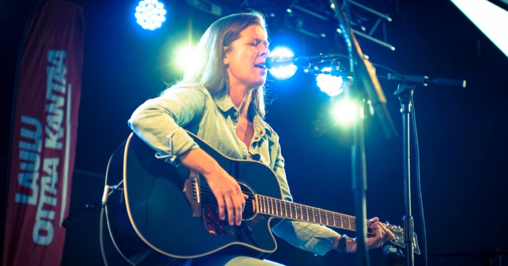 Henkilö soittaa kitaraa ja laulaa mikrofoniin esiintymislavalla. Taustalla näkyy lippu, jossa lukee Laulu ottaa kantaa.