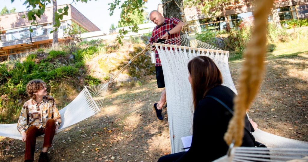 Kaksi henkilöä istuu omissa riippumatoissaan hotellin takana olevassa metsikössä. He juttelevat kolmannen, seisovan henkilön kanssa.
